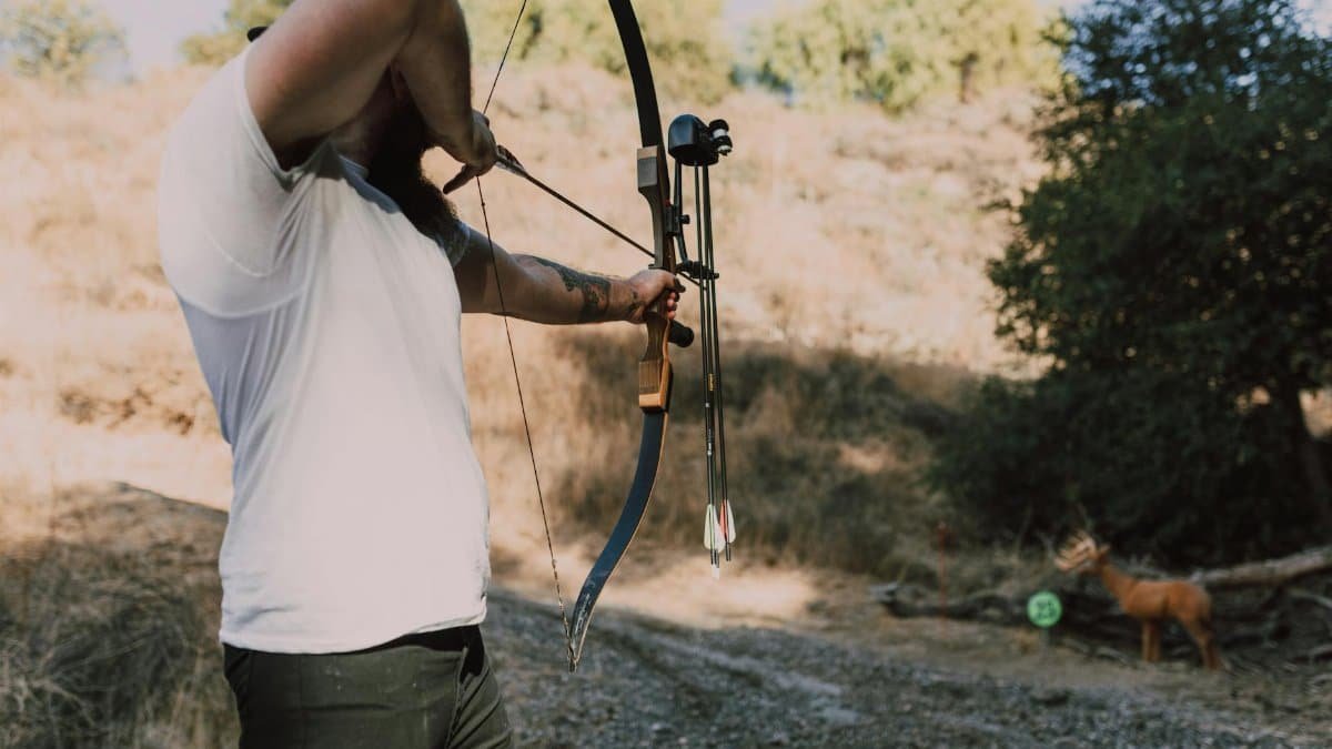 An adult man practices archery with a bow and arrow in a natural outdoor setting.
