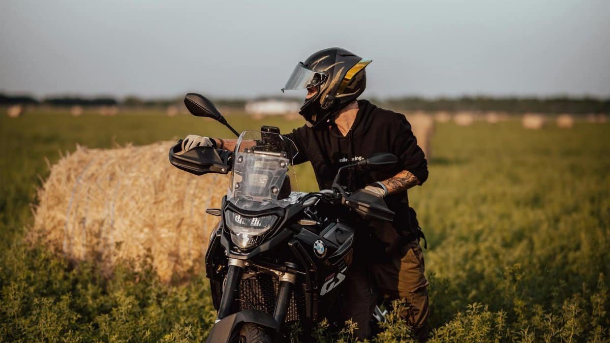 A motorcyclist pauses near a hay bale in a serene open field, enjoying the sunset.
