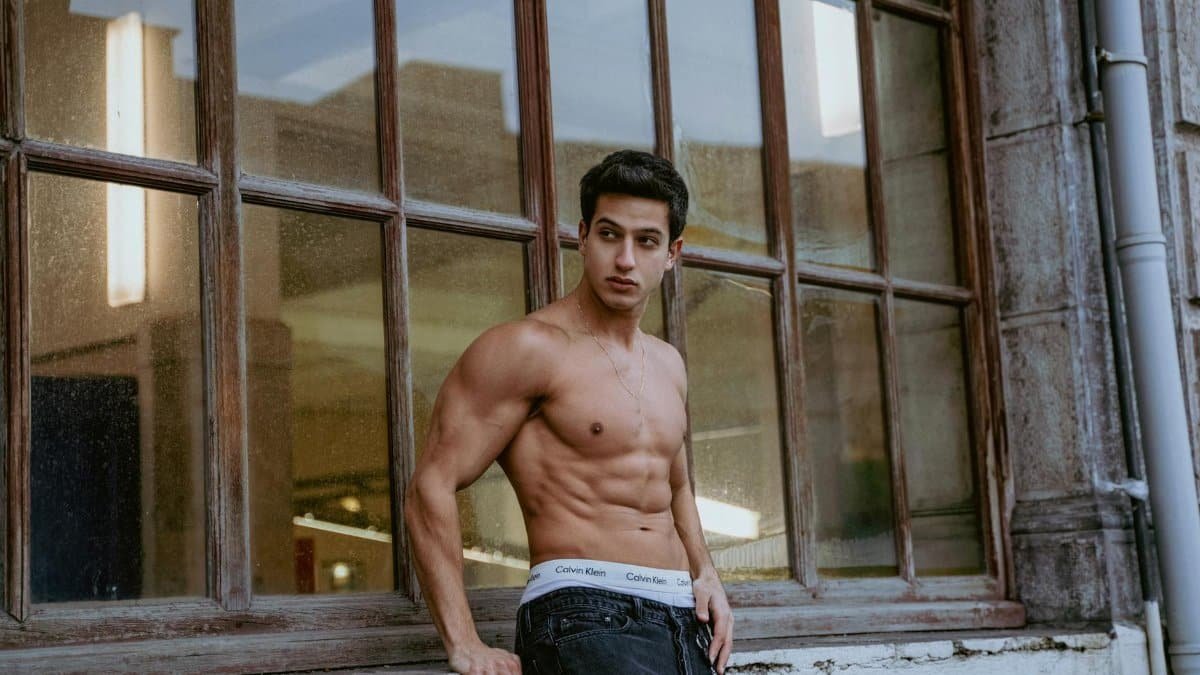 A muscular shirtless man models near a large window in Istanbul, showcasing urban style and fitness.