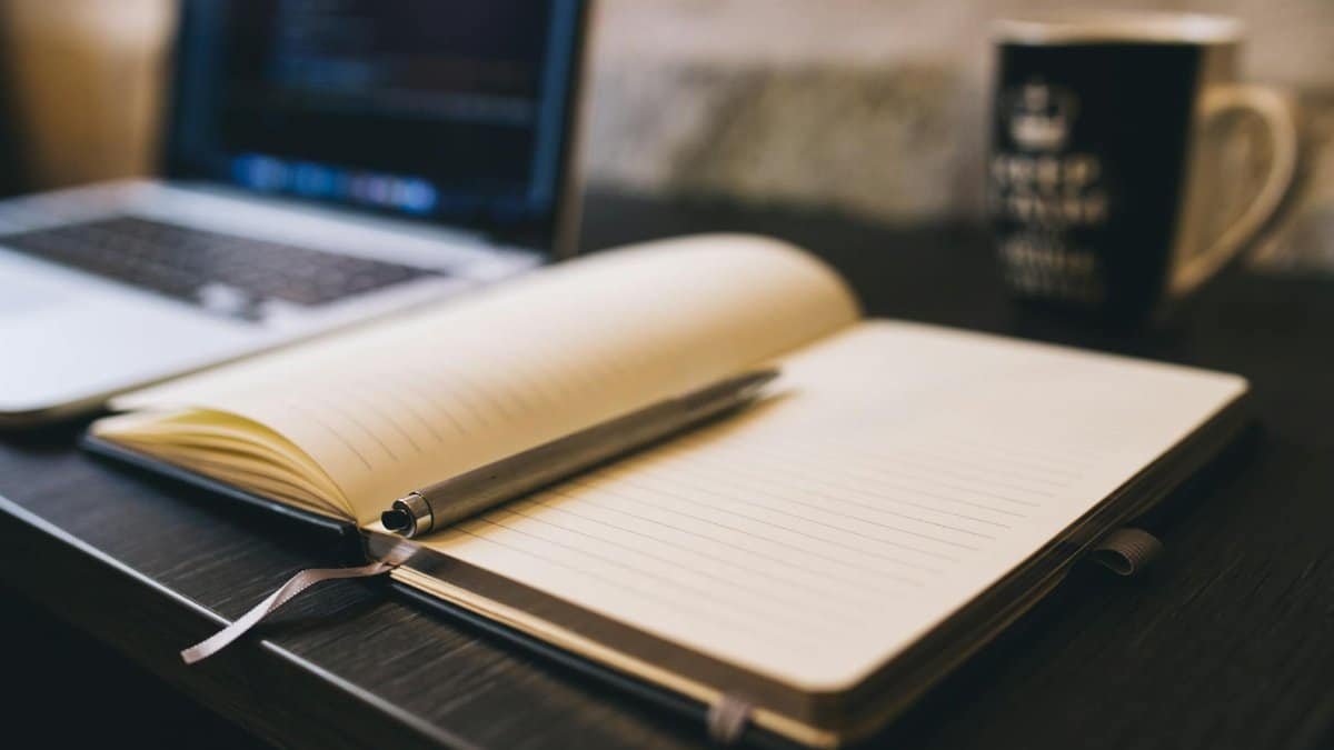 Open notebook with a pen on a wooden desk beside a laptop, ideal for blogging and work-related themes.