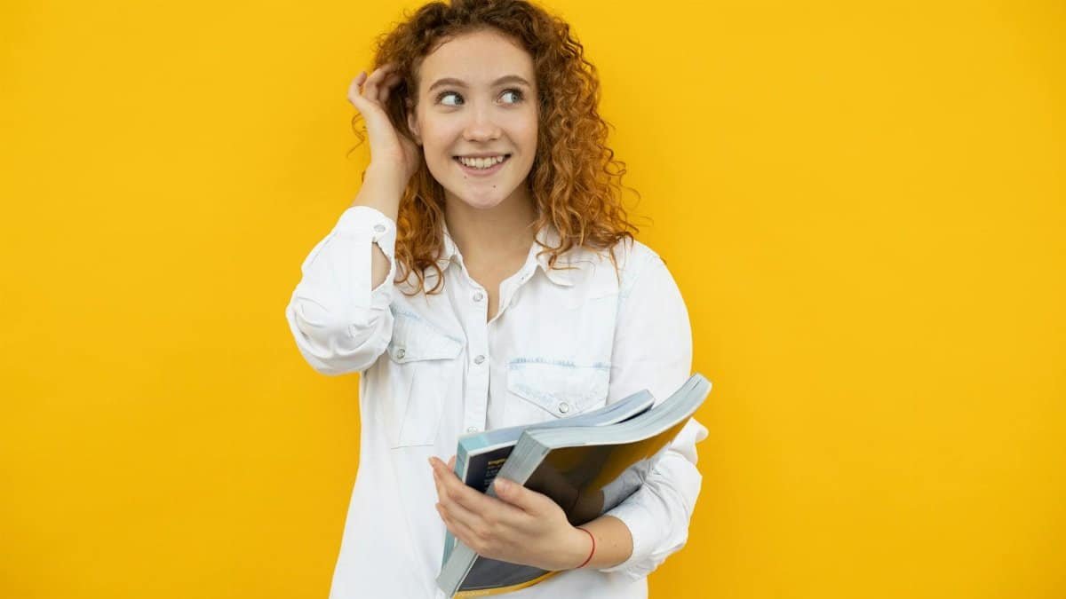 Cheerful young woman standing with books in hand against a vibrant yellow backdrop.