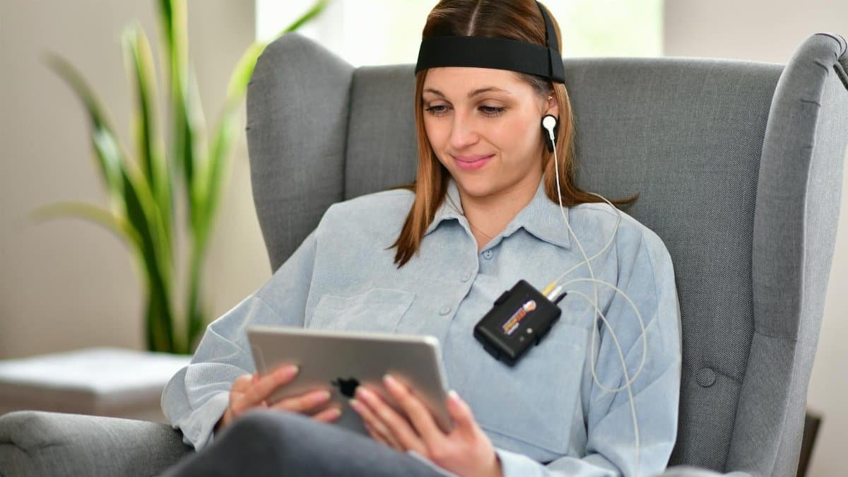 Woman using neurofeedback device on armchair, engaging with technology indoors.