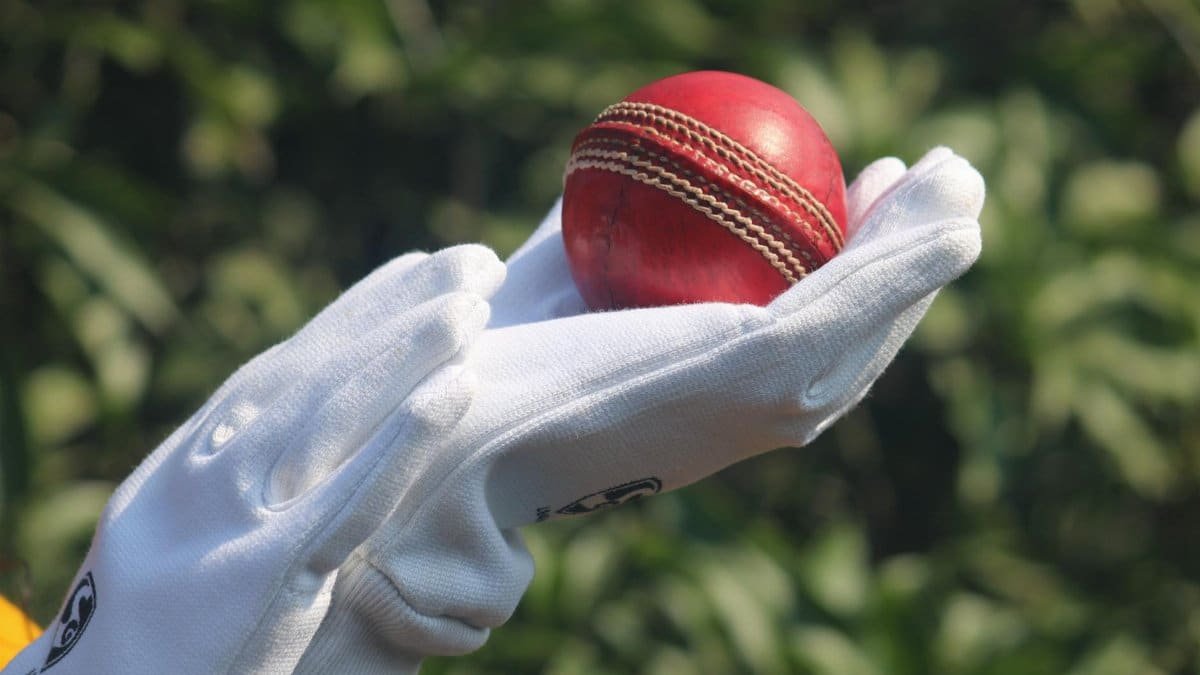 Detailed photo of a red cricket ball held by gloved hands outdoors.