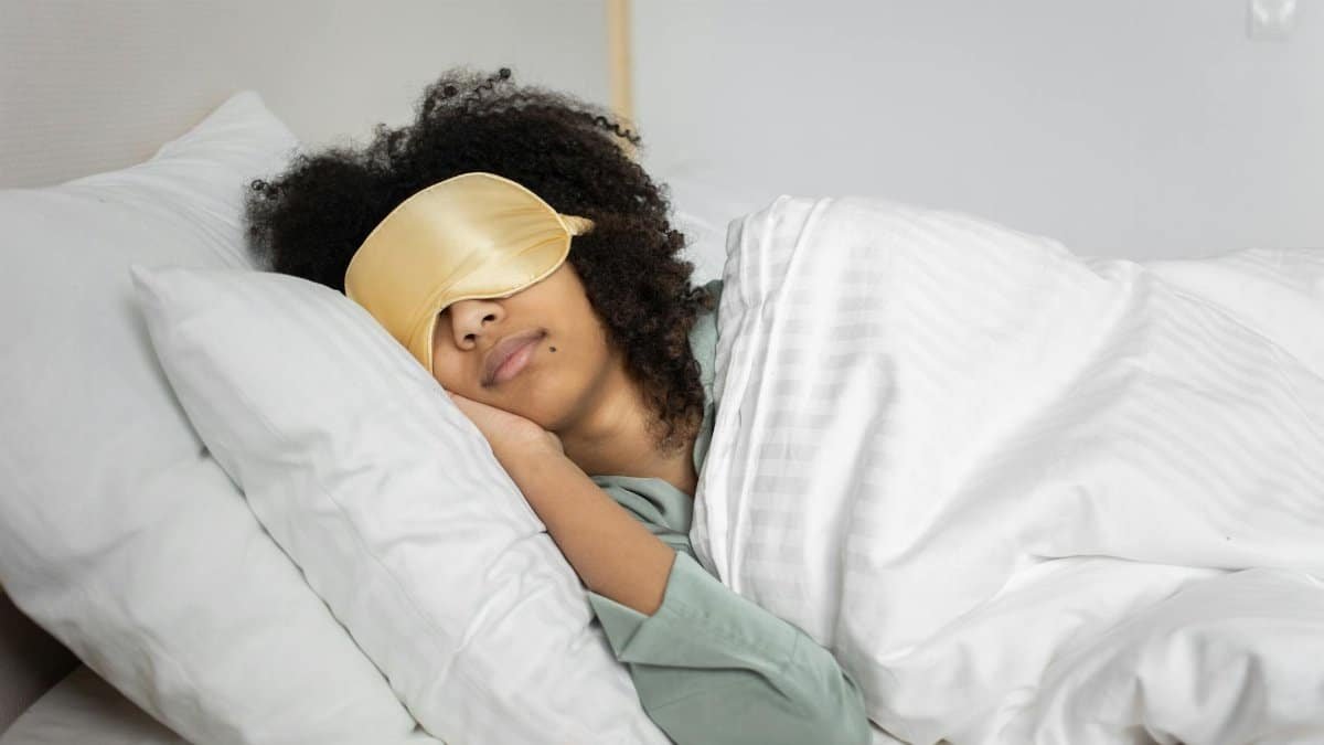 A woman with afro hair sleeps soundly in bed with a sleep mask, enjoying a cozy indoor atmosphere.