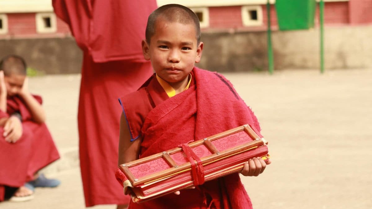 A young Buddhist monk in traditional clothing holding Buddhist scriptures outdoors.
