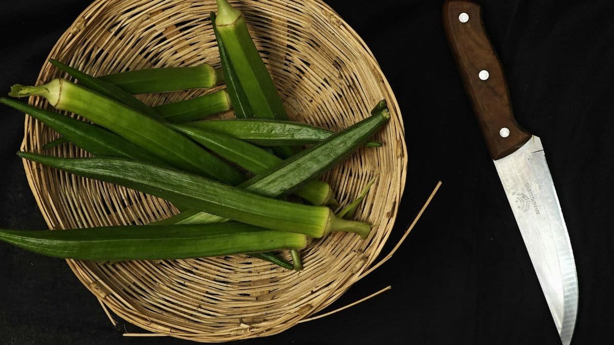 Top view of fresh okra in a woven basket beside a wooden-handled kitchen knife.