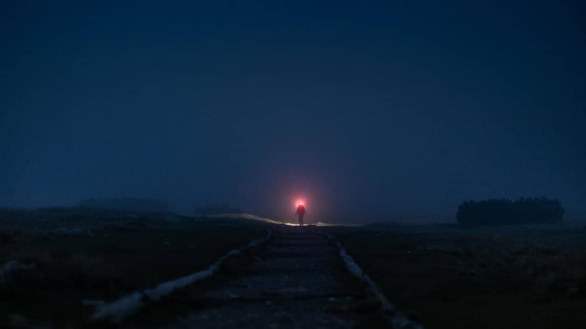 A person with a flashlight walking on a foggy path at night, creating an eerie and adventurous atmosphere.