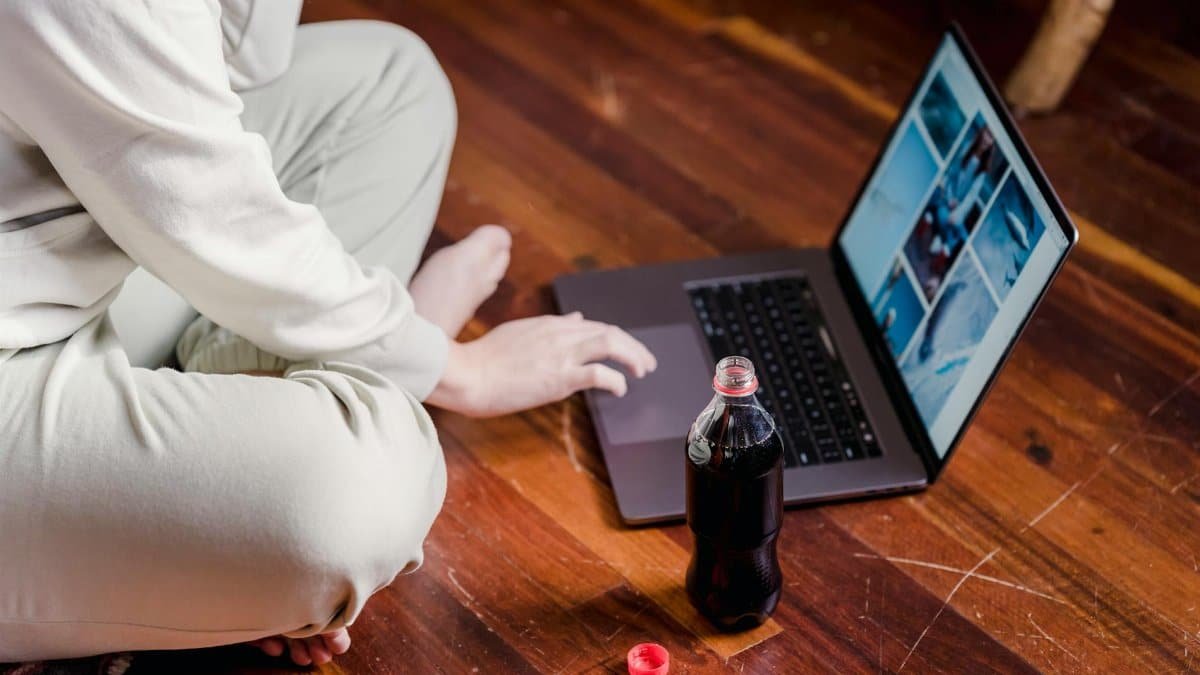 Side view of unrecognizable barefoot person with crossed legs browsing internet on netbook while sitting on floor with bottle of soda at home