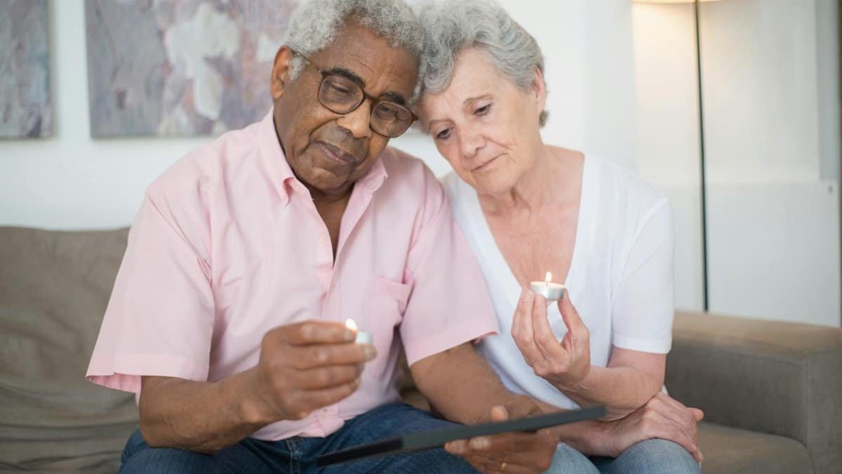 A grieving elderly couple holding candles, reminiscing over a photograph indoors.