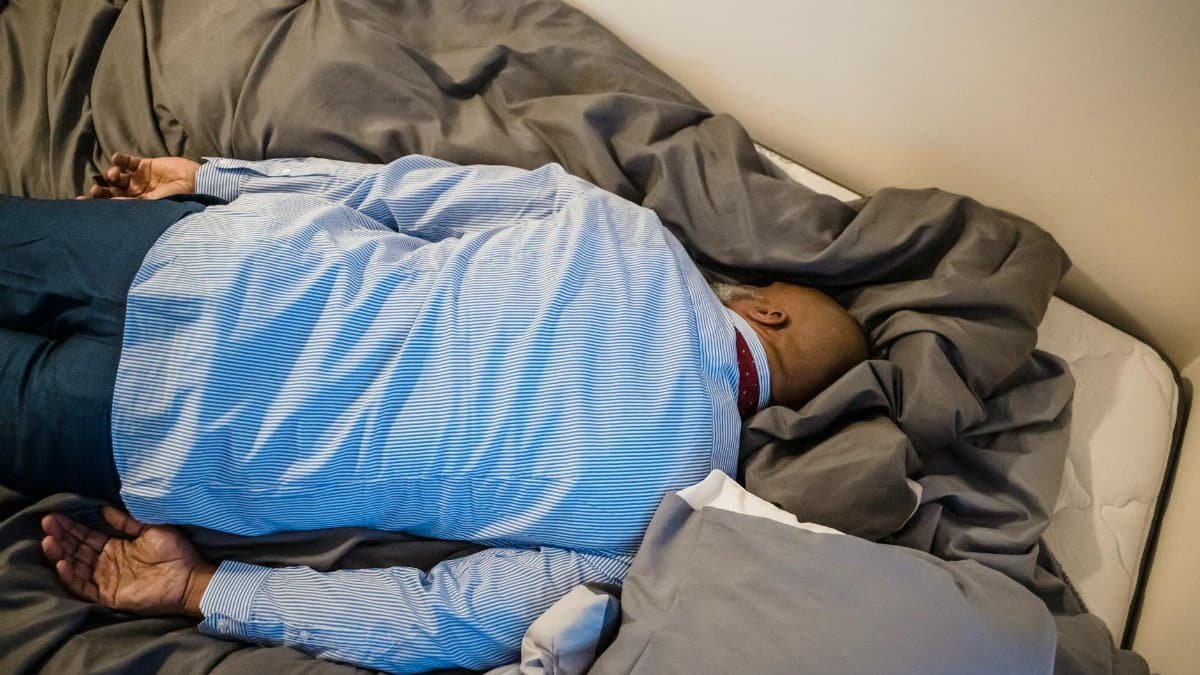 A man in formal attire rests face down on a bed, conveying exhaustion and fatigue.