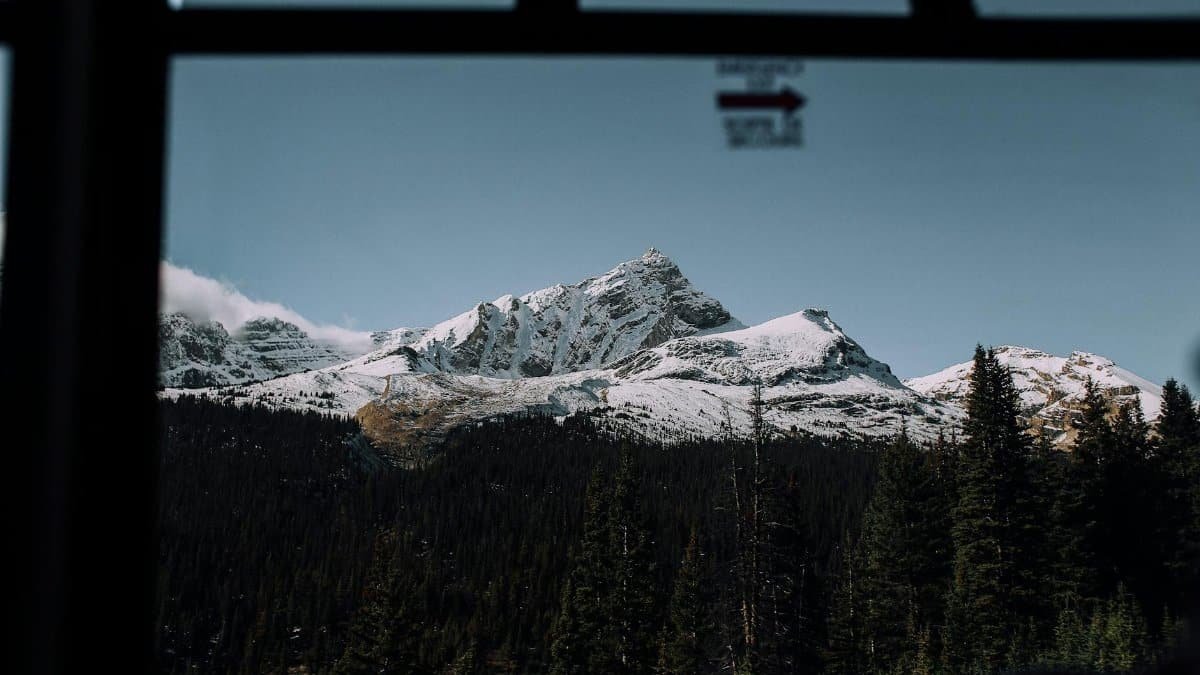 Through glass view of high mountains covered with snow near green woods under blue cloudless sky in nature