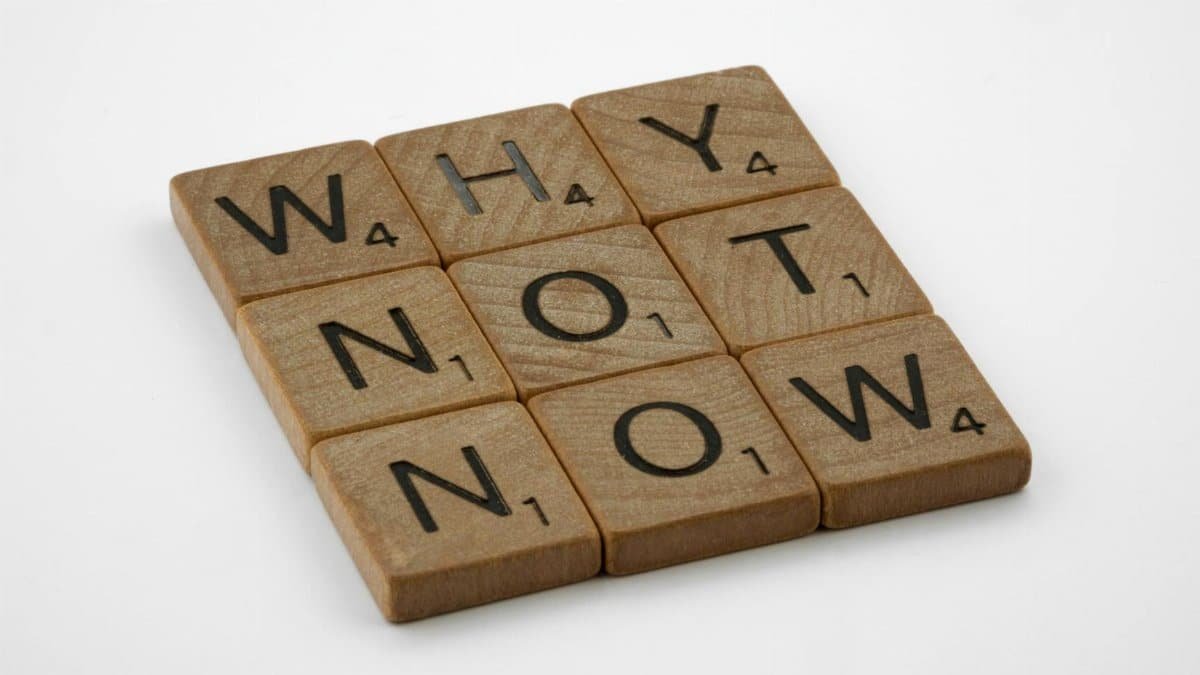 Wooden letter tiles form the motivating phrase 'Why Not Now' on a white background, encouraging action and decision-making.