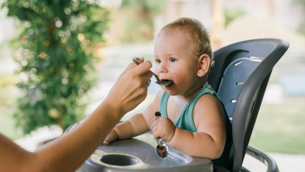 Cute baby eating with a spoon in a high chair outdoors. Calm and precious moment.