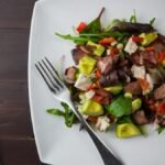 A fresh bacon, avocado, and mixed greens salad served on a square plate with a fork.