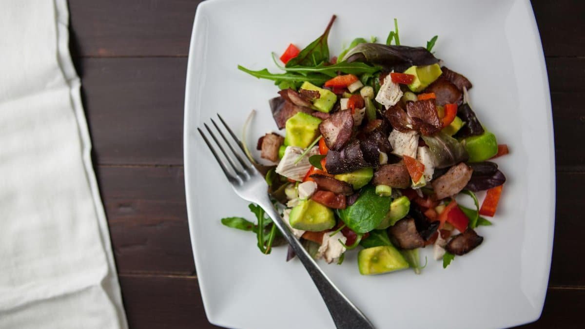 A fresh bacon, avocado, and mixed greens salad served on a square plate with a fork.