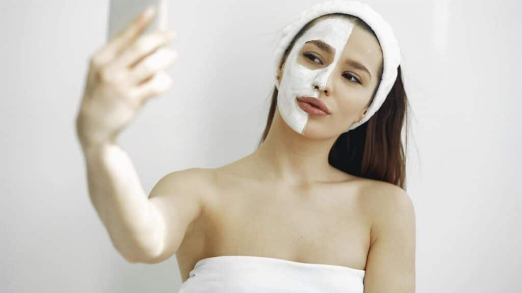 Young woman with a facial mask on half her face taking a selfie indoors. Beauty and skincare concept.