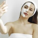 Young woman with a facial mask on half her face taking a selfie indoors. Beauty and skincare concept.