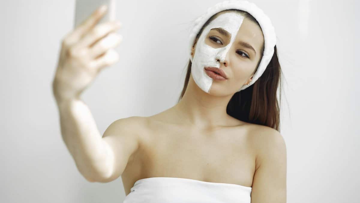 Young woman with a facial mask on half her face taking a selfie indoors. Beauty and skincare concept.