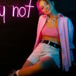 Young woman posing indoors under pink neon 'why not' sign, expressing individuality.