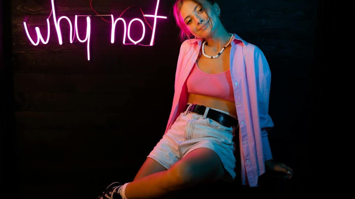 Young woman posing indoors under pink neon 'why not' sign, expressing individuality.