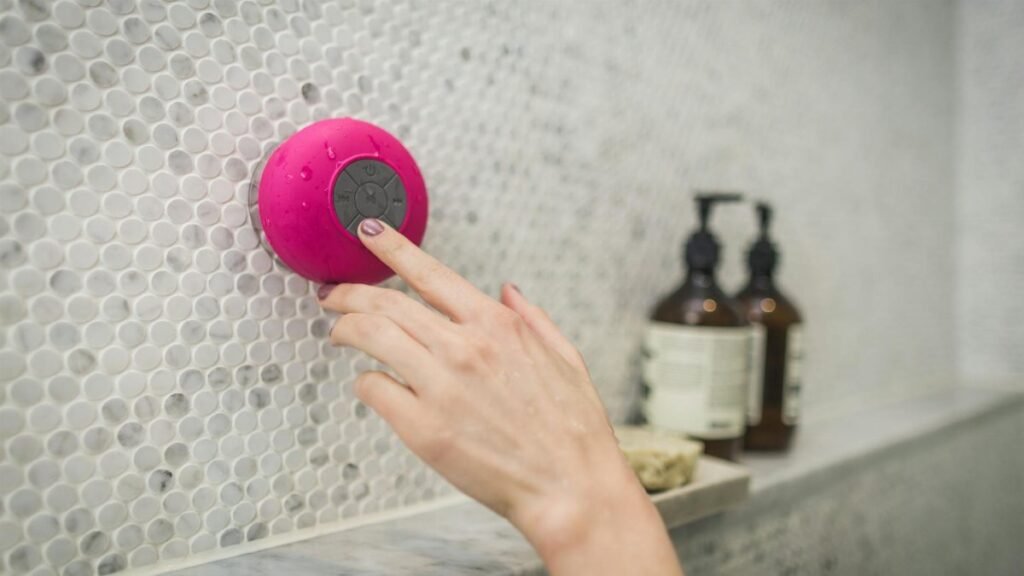 A person adjusting a pink waterproof bluetooth speaker in a tiled bathroom setting.