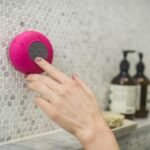 A person adjusting a pink waterproof bluetooth speaker in a tiled bathroom setting.