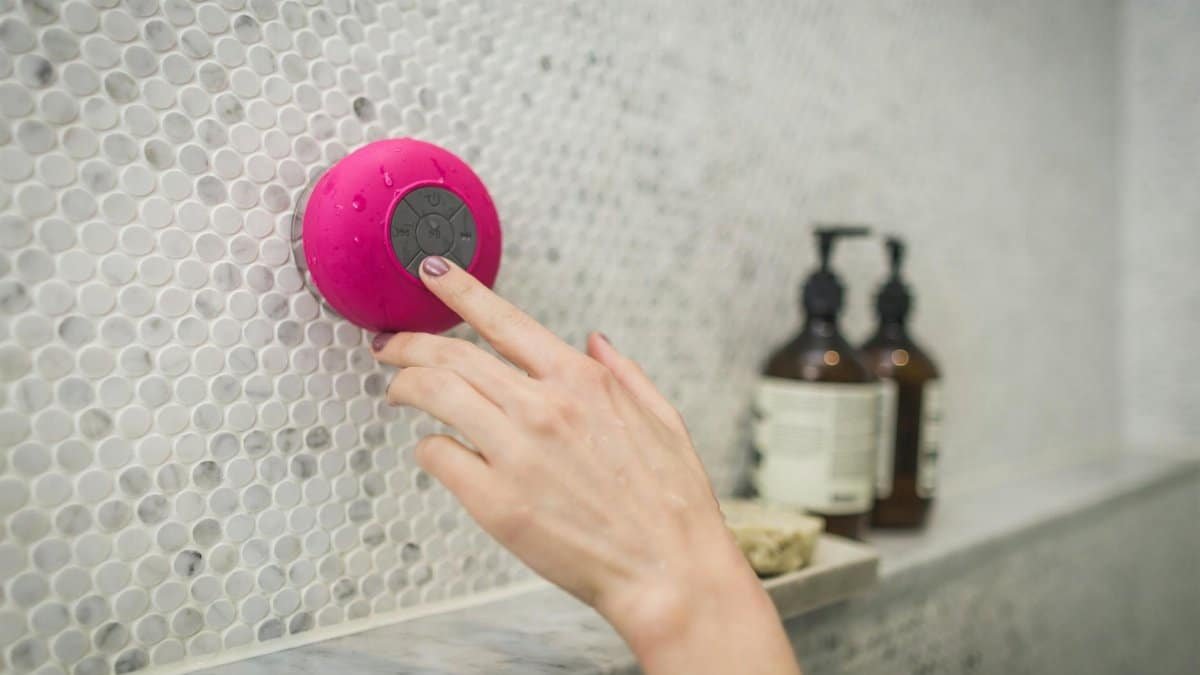 A person adjusting a pink waterproof bluetooth speaker in a tiled bathroom setting.