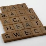 Wooden Scrabble tiles arranged to form the words FAT, SALT, ACID, HEAT, and SWEET on a white surface.