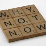 Wooden letter tiles form the motivating phrase 'Why Not Now' on a white background, encouraging action and decision-making.