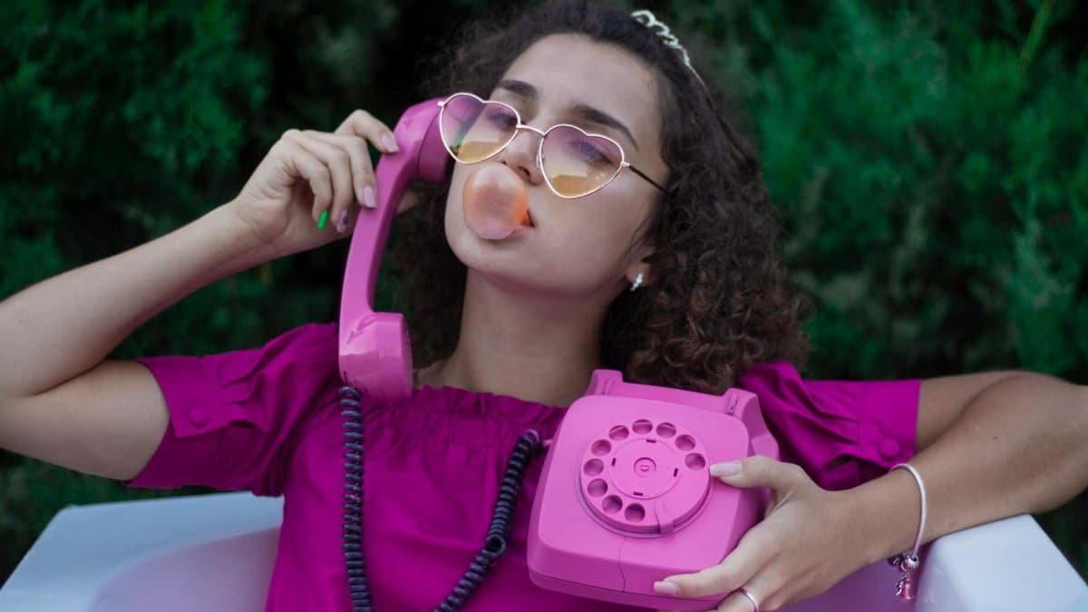 Chic woman with curly hair, blowing bubble gum and holding a pink retro telephone outdoors.