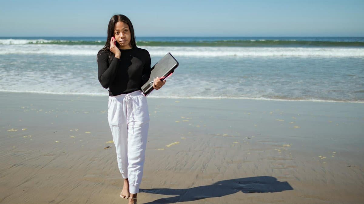 A woman works remotely on the beach, balancing a phone call with her laptop.