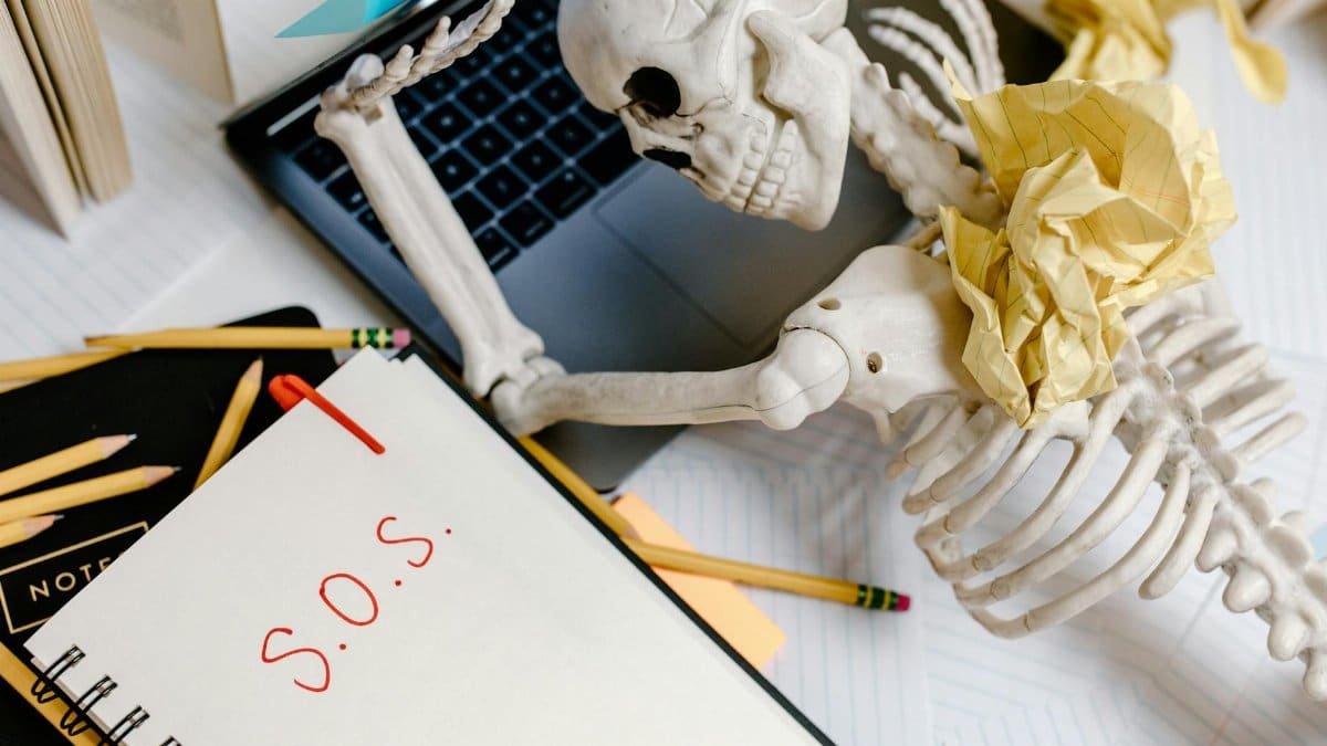 A humorous depiction of office burnout with a skeleton and S.O.S note at a cluttered desk.