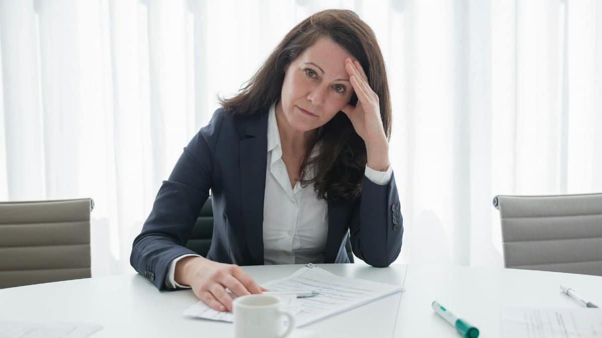 A stressed businesswoman in corporate attire sits at a desk with papers, looking overwhelmed.