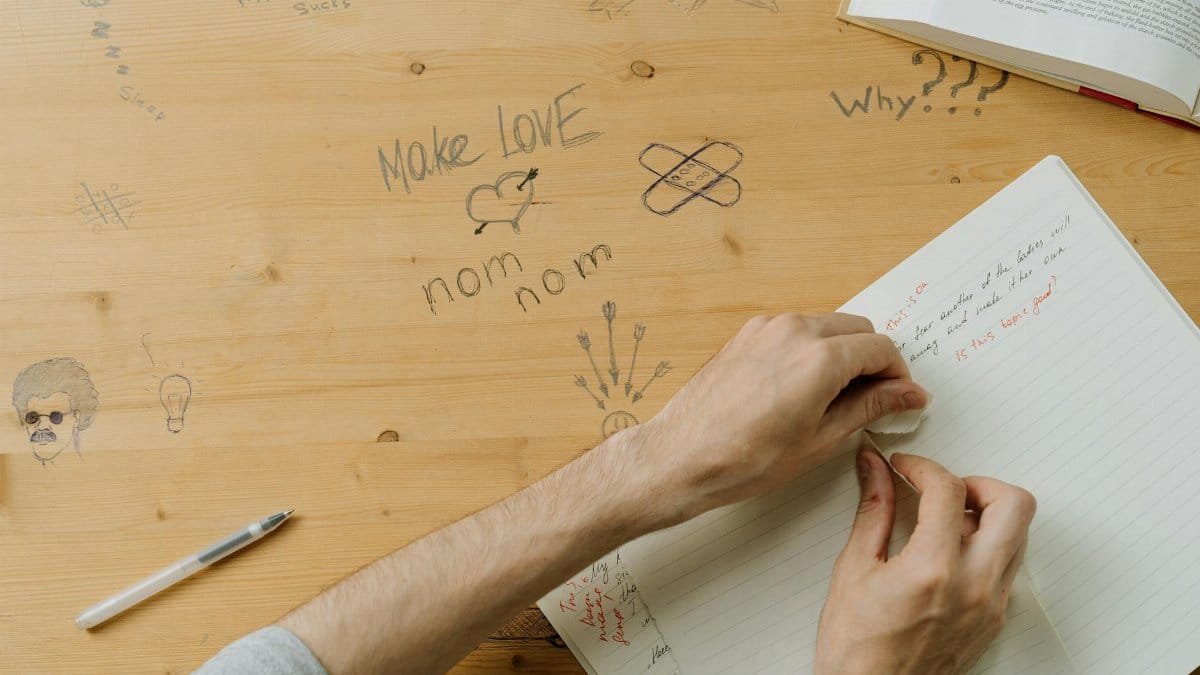 A person writing notes in a notebook on a doodle-covered wooden desk.