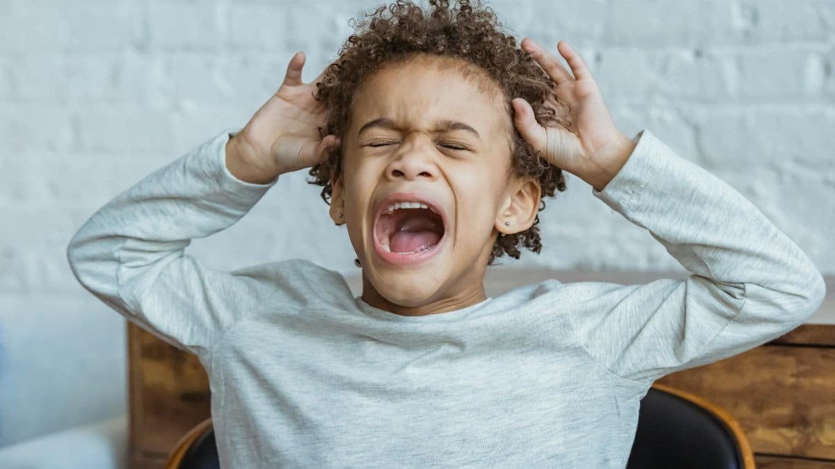 Young boy showing frustration indoors with hands on head, eyes closed, mouth open.
