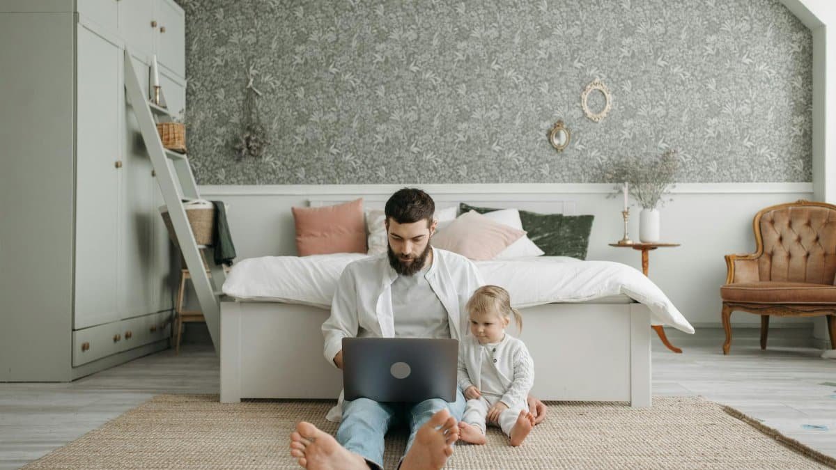 A bearded father works on a laptop while sitting on the bedroom floor with his young daughter, embracing a remote work lifestyle.