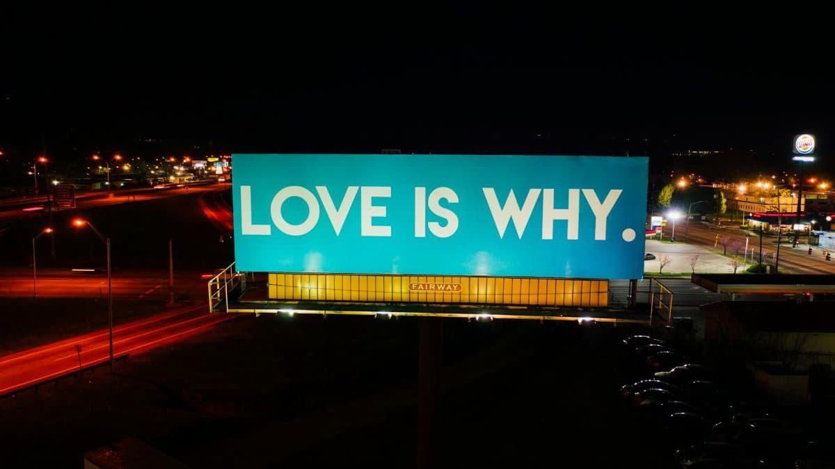 Blue billboard saying Love is why placed on road surrounded by cars and street lights against black night