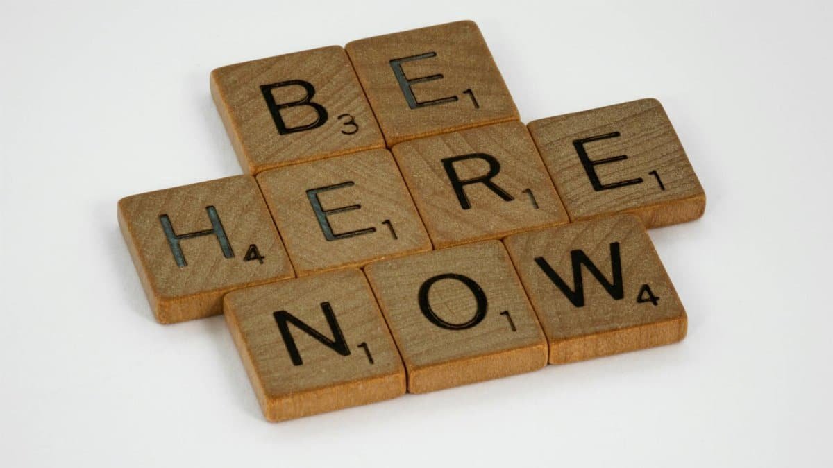 Inspirational text 'Be Here Now' created with wooden Scrabble tiles on white background.