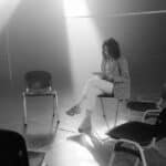 A solitary woman sits in a smoky, sunlit room surrounded by empty chairs, conveying a sense of solitude.