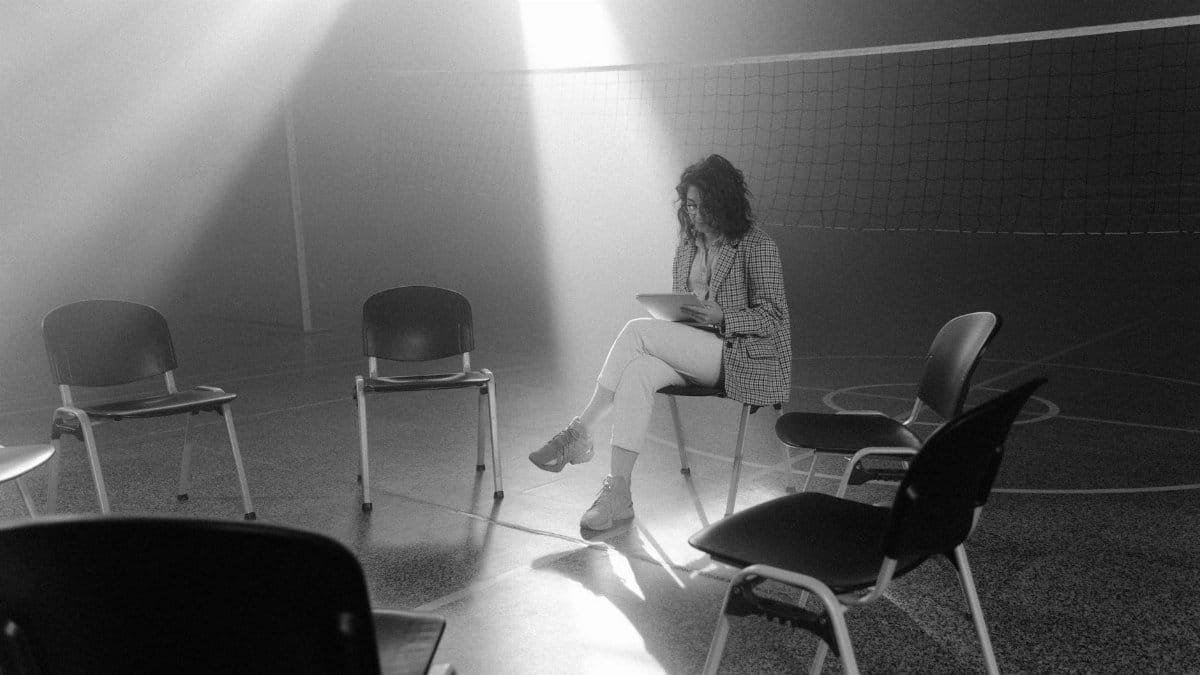 A solitary woman sits in a smoky, sunlit room surrounded by empty chairs, conveying a sense of solitude.