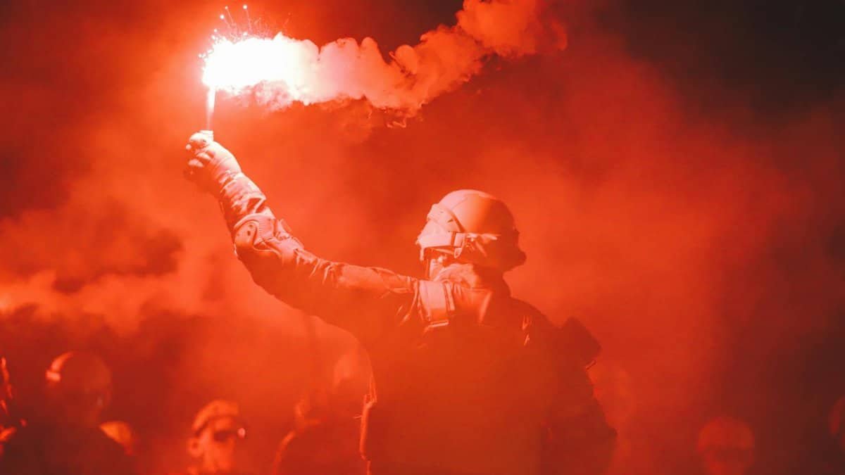A soldier signals with a red flare, surrounded by dense smoke and darkness, creating a dramatic scene.