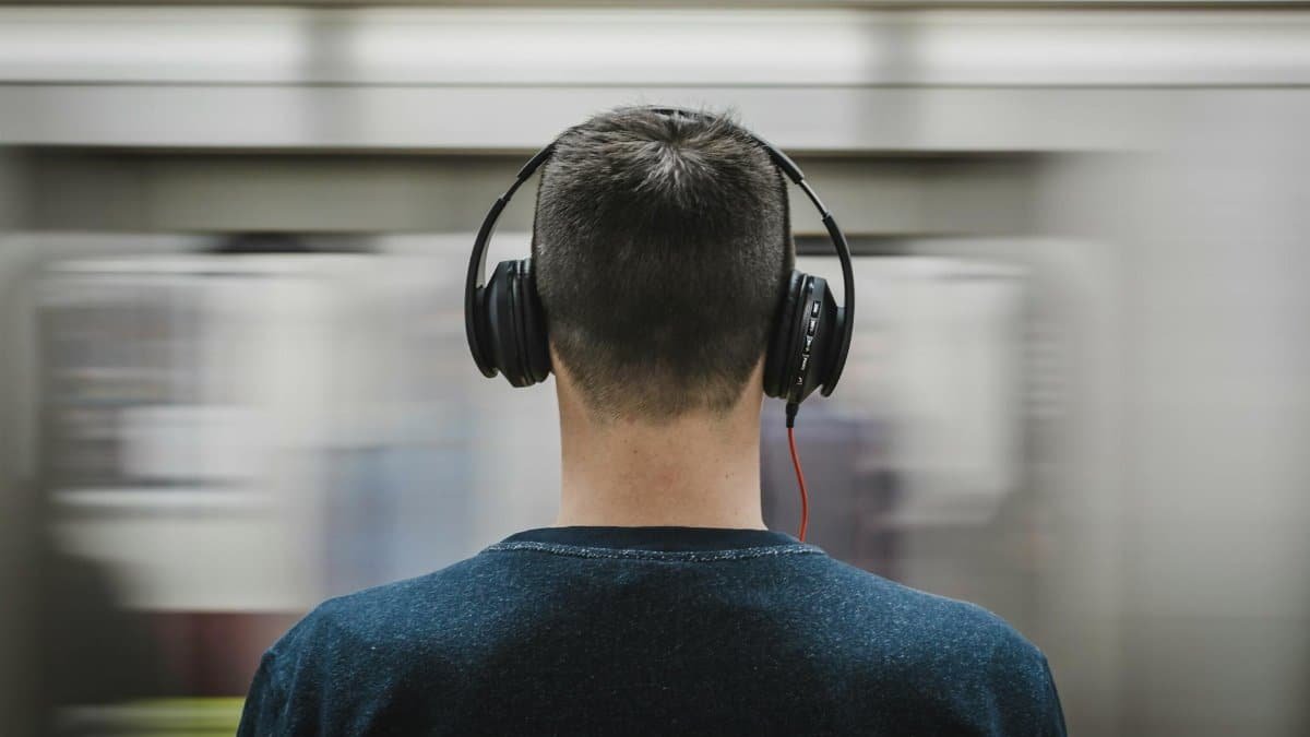Rear view of a man with headphones standing at a subway platform, blurred train passing by.