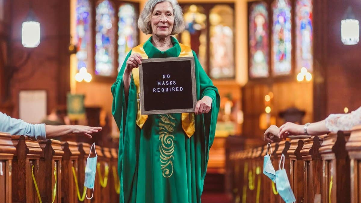 Elderly female priest in church holding a 'No masks required' sign, symbolizing post-pandemic life.