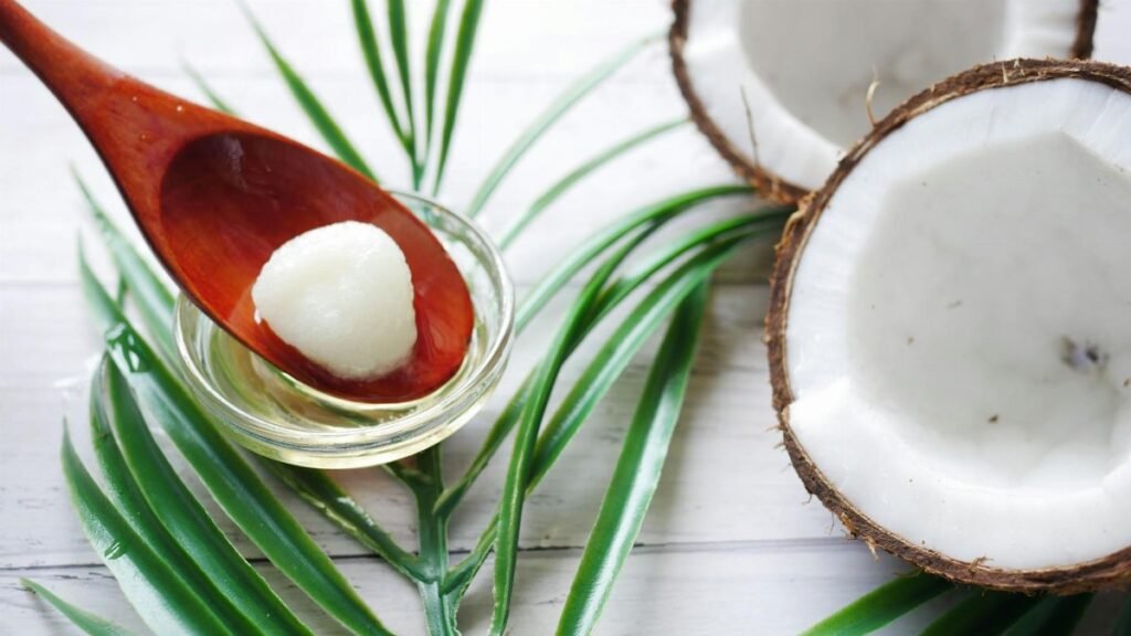 A close-up of organic coconut oil on a wooden spoon beside fresh coconut and palm leaves.