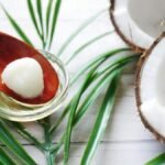 A close-up of organic coconut oil on a wooden spoon beside fresh coconut and palm leaves.