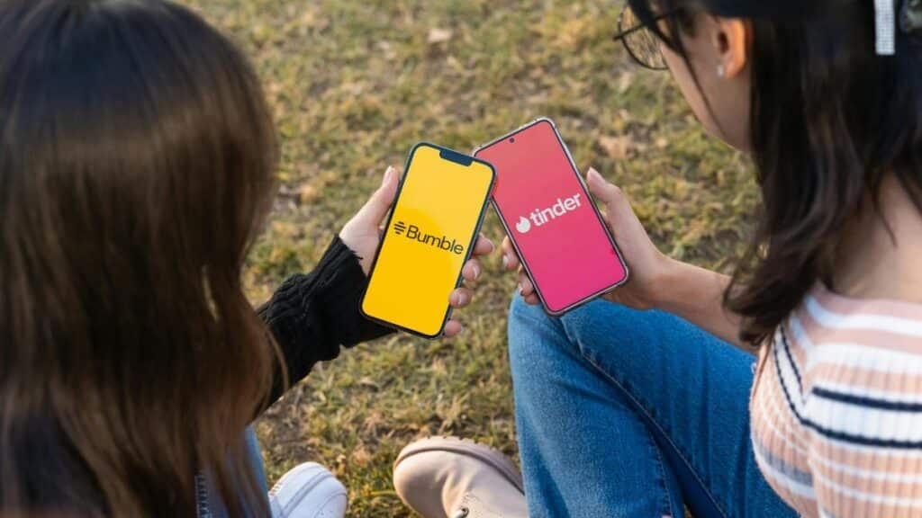 Two women using Bumble and Tinder apps on smartphones in a park setting.