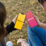 Two women using Bumble and Tinder apps on smartphones in a park setting.