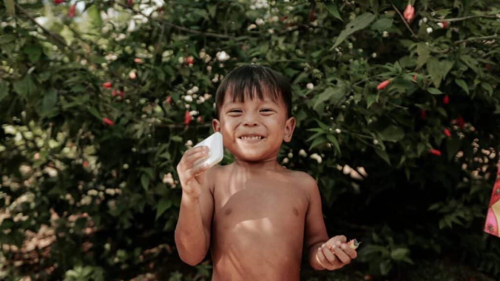 A smiling child holding a bar of soap outdoors among lush greenery, evoking a sense of cleanliness and happiness.