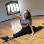 Young woman in activewear taking a selfie while doing the splits on a wooden floor.