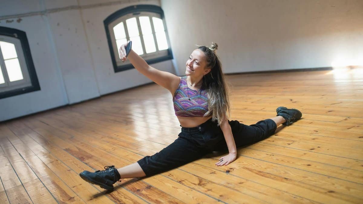 Young woman in activewear taking a selfie while doing the splits on a wooden floor.